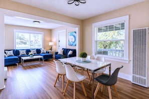 A modern living room with a sofa set, dining table, and chairs, large windows with blinds, and a ceiling fan. - CasaViva Vista · Top-Floor 2BR with Private Porch, City Views, Walk to Eats (San Diego)