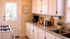 A kitchen with white cabinets, a stove, a trash can, a coffee maker, and a sign on the wall. - CasaViva Breeze 2BR w/ Sunset Views, Firepit & Backyard — Walk to Fun & Eats (San Diego)