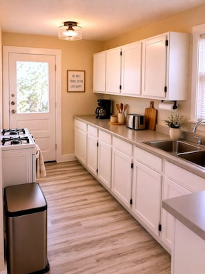 A kitchen with white cabinets, a stove, a trash can, a coffee maker, and a sign on the wall. - CasaViva Breeze 2BR w/ Sunset Views, Firepit & Backyard — Walk to Fun & Eats (San Diego)