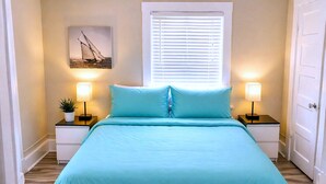 A bed with a teal bedspread, two bedside tables with lamps, a window with blinds, and a wall-mounted picture. - CasaViva Breeze 2BR w/ Sunset Views, Firepit & Backyard — Walk to Fun & Eats (San Diego)