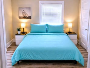 A bed with a teal bedspread, two bedside tables with lamps, a window with blinds, and a wall-mounted picture. - CasaViva Breeze 2BR w/ Sunset Views, Firepit & Backyard — Walk to Fun & Eats (San Diego)