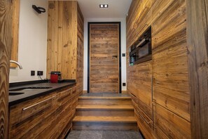 Un couloir étroit aux murs recouverts de lambris en bois, un comptoir noir sur lequel repose un appareil électroménager rouge, et une porte au fond.