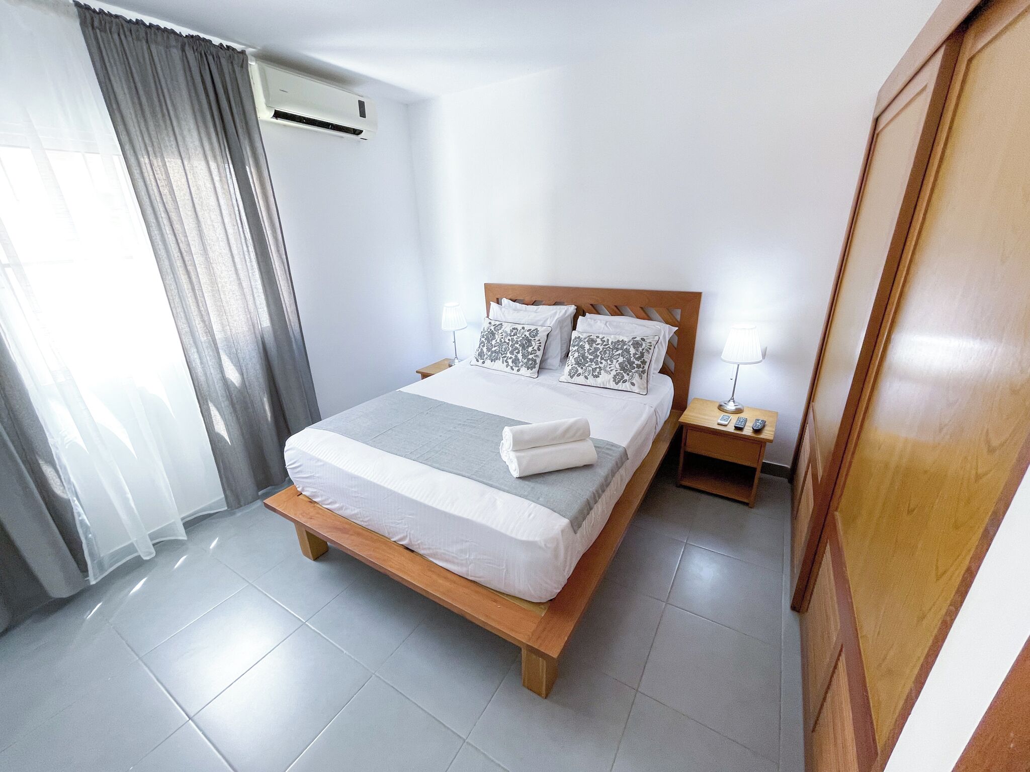 Una habitación de hotel con una cama de madera, una mesita de noche, una lámpara y un ventanal con cortinas.
