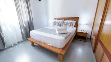 Ein ordentlich bezogenes Bett mit Holzrahmen, ein Nachttisch mit Lampe und ein Fenster mit Vorhängen.