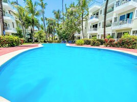 Una piscina rodeada de edificios de varios pisos con balcones, palmeras y jardines bien cuidados.