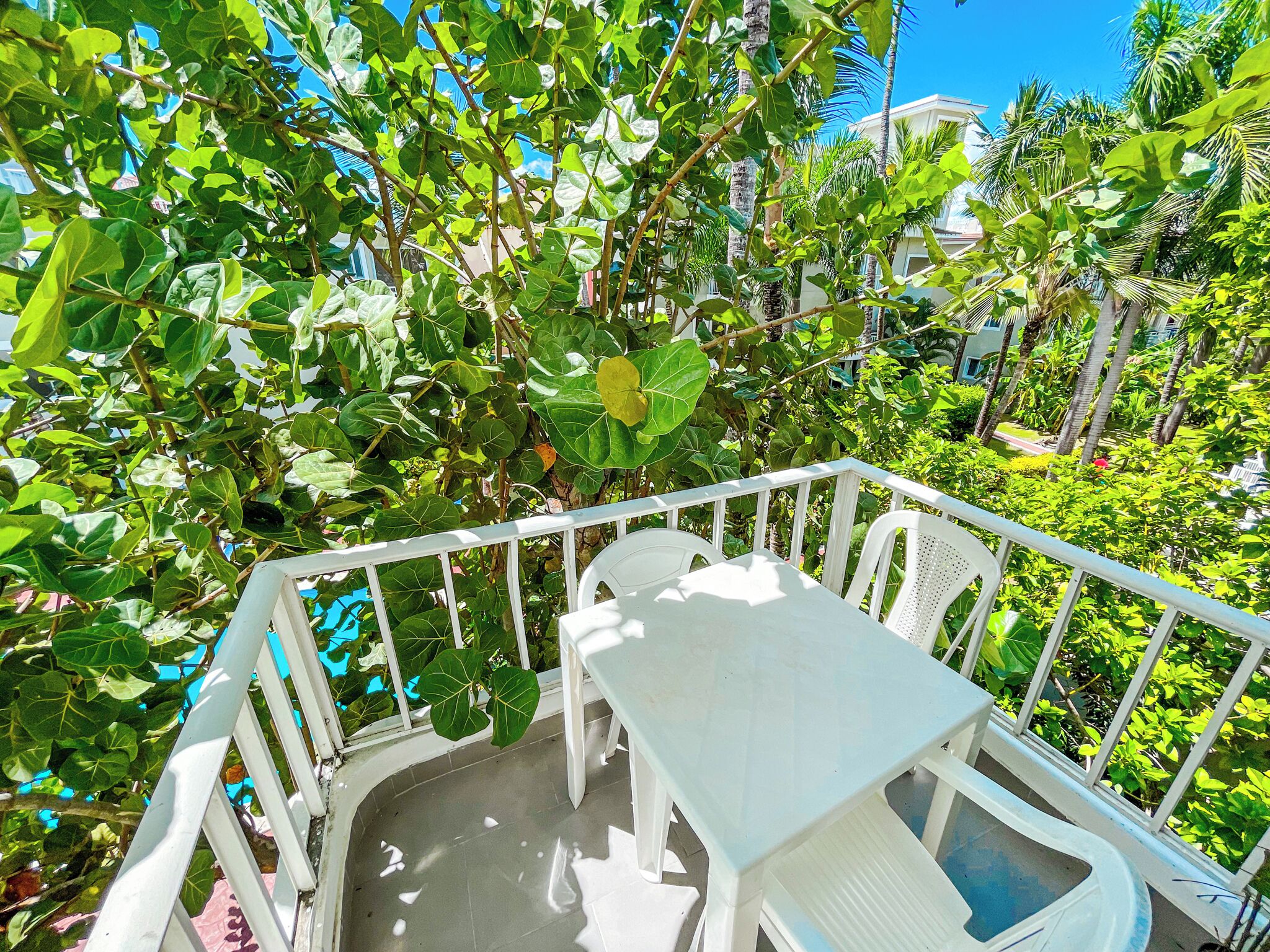 Un balcon avec des balustrades blanches, une table blanche et des chaises, le tout entouré d’un luxuriant feuillage verdoyant.