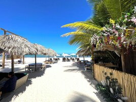 Zona de playa con sombrillas de paja, sillas de descanso y una valla de madera decorada con plantas.