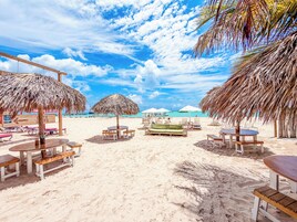 Zona de playa con sombrillas de paja, mesas y bancos de madera.