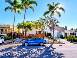 Een woonstraat met huizen, een blauwe auto aan de kant van de weg en palmbomen.