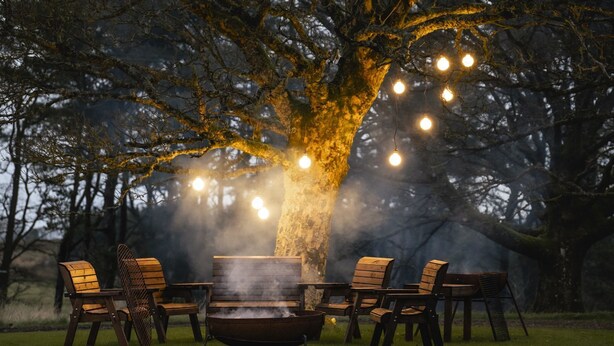 A rustic outdoor seating area with a fire pit, surrounded by chairs and illuminated by string lights.