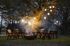 Un espace extérieur rustique aménagé avec des sièges, un foyer et entouré de chaises, le tout illuminé par des guirlandes lumineuses.