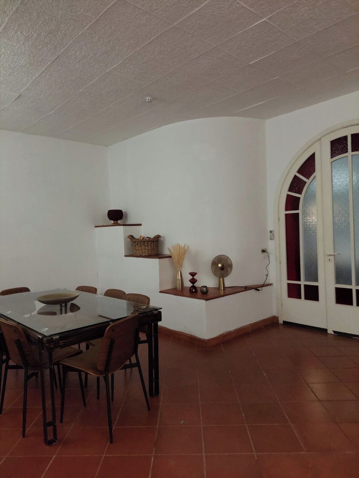 A dining area with a glass-top table and chairs, a small shelf with decorative items, and a door with frosted glass panels.
