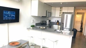 Una cocina moderna con armarios blancos, electrodomésticos de acero inoxidable, un televisor de pantalla plana y una zona de bar con taburetes.