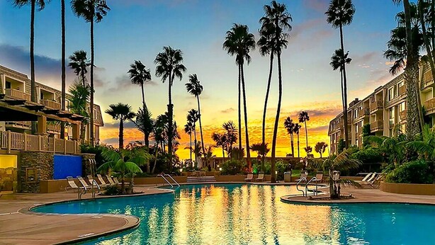 Une piscine d’un complexe hôtelier, avec des palmiers et un coucher de soleil en arrière-plan.