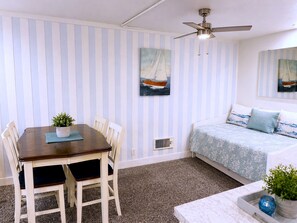 Ein Zimmer mit Esstisch, Stühlen, Deckenventilator, einem Bett mit gemustertem Überwurf und zwei Wandbildern.