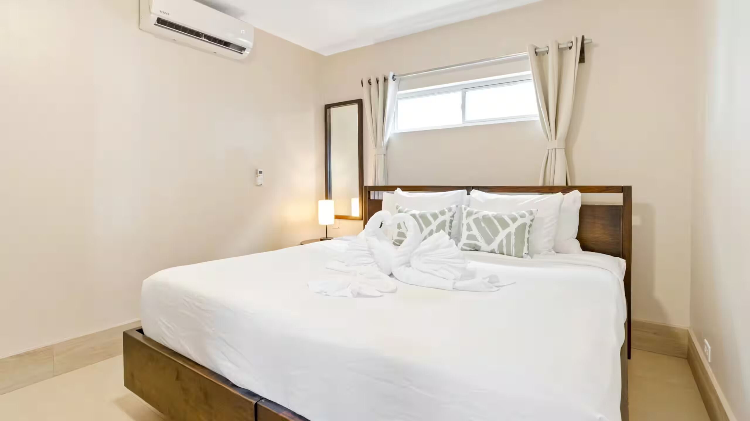 A hotel room with a bed, a nightstand, a lamp, a window with curtains, and an air conditioning unit.