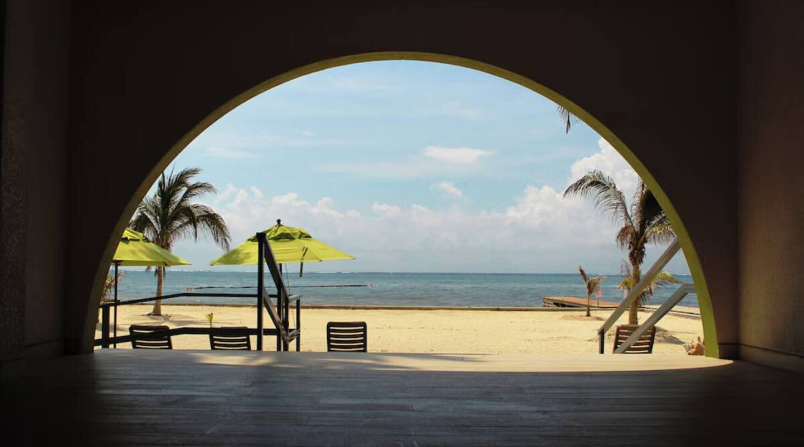 A beach view through an arched window with a table and chairs, palm trees, and a clear sky.