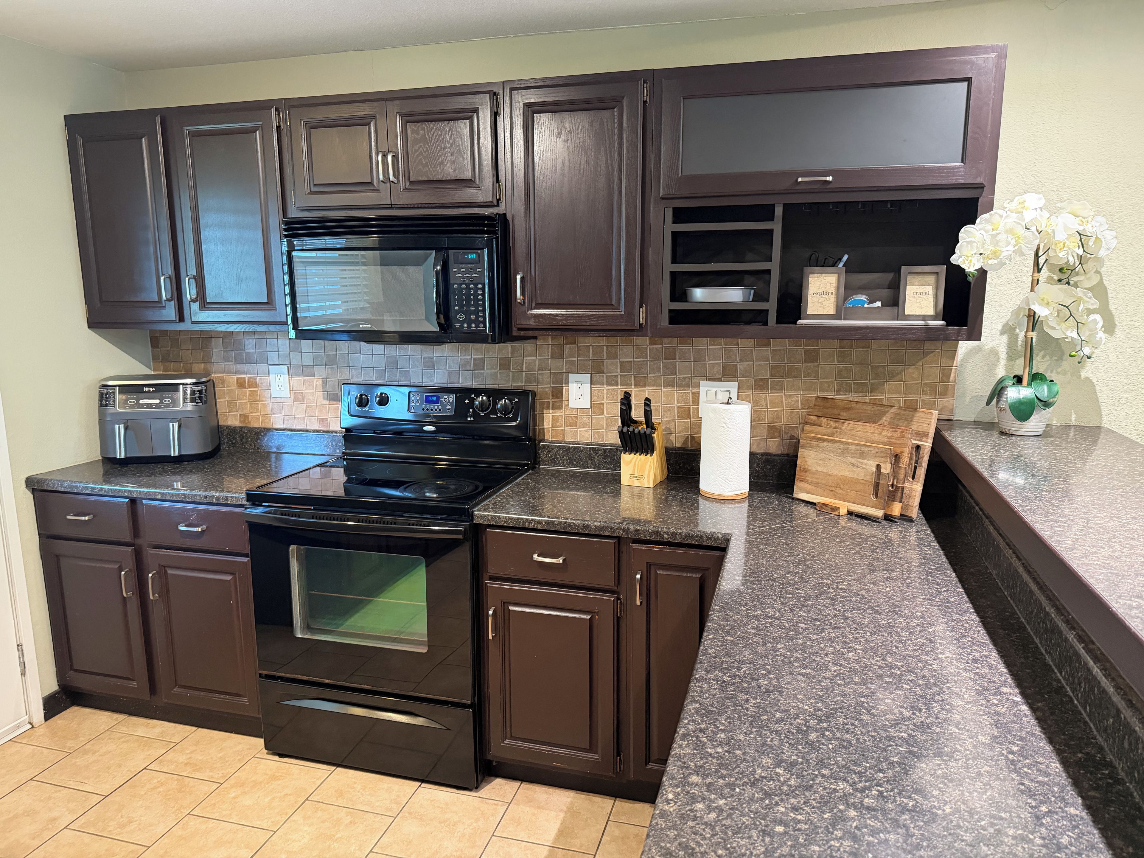 A kitchen with dark brown cabinets, a black microwave, a stove, and a countertop with a knife block and a vase of flowers.