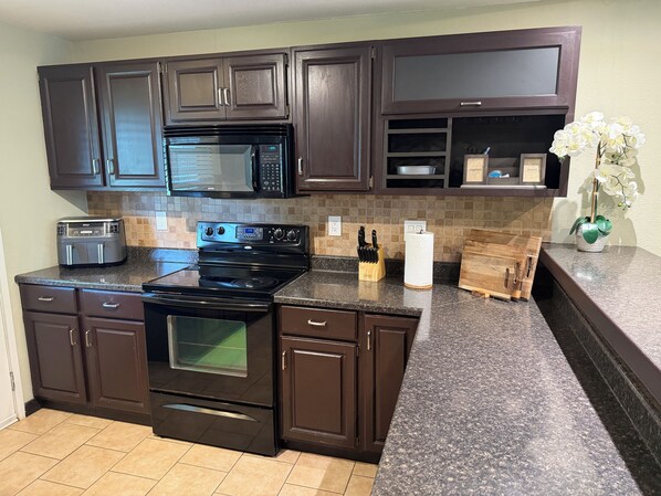 A kitchen with dark brown cabinets, a black microwave, a stove, and a countertop with a knife block and a vase of flowers.