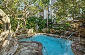 Una piscina rodeada de formaciones rocosas naturales y árboles, con un edificio al fondo.