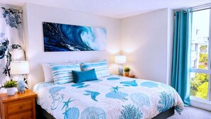A bedroom with a bed, nightstands, a wall painting, and a window with curtains. - SolMar Vista 2BR/2BA Oceanview, Heated Pools, Hot Tubs & Steps to Beach (Oceanside)