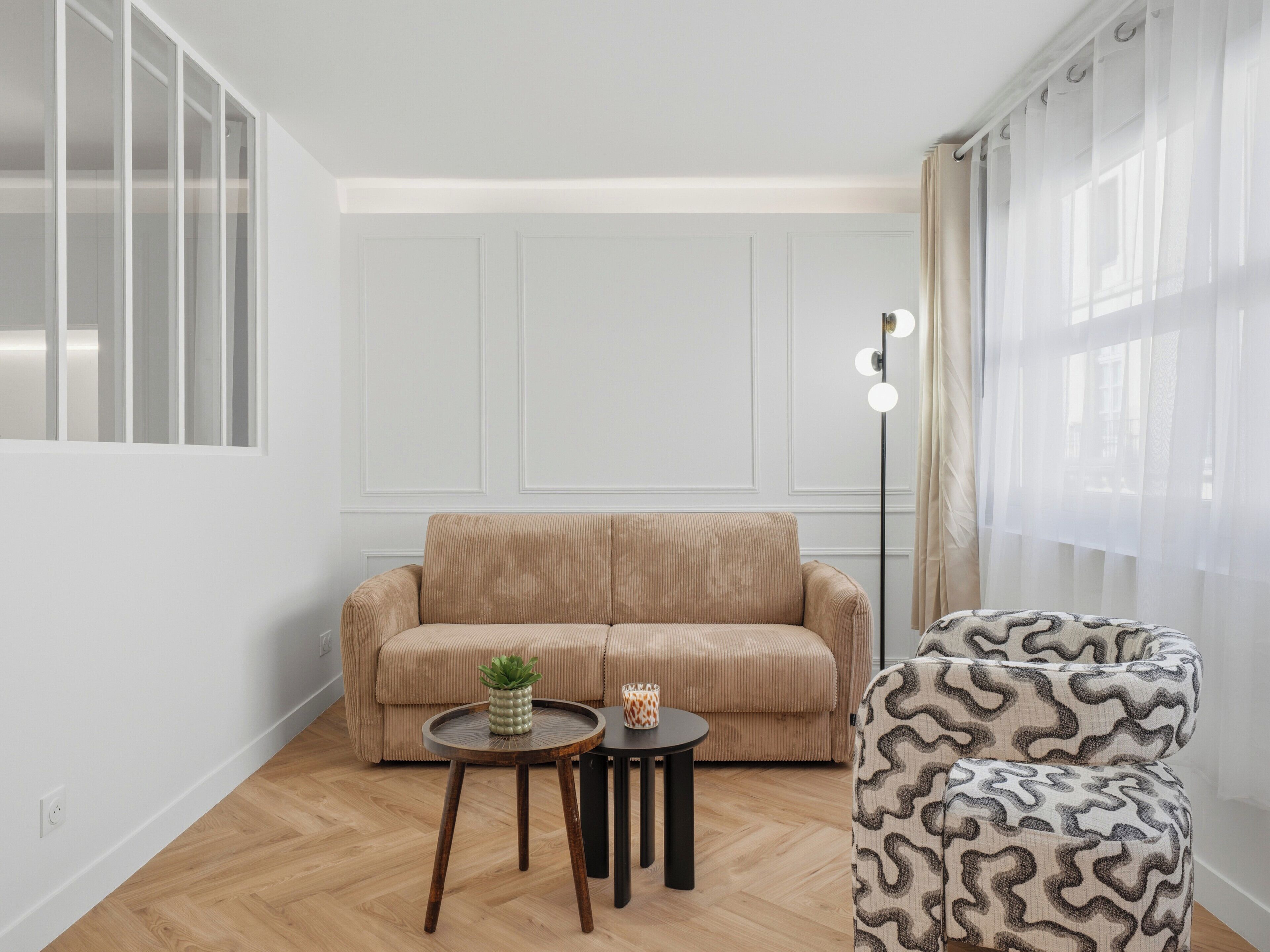 Uma sala de estar com sofá bege, poltrona estampada, duas mesinhas e um abajur.