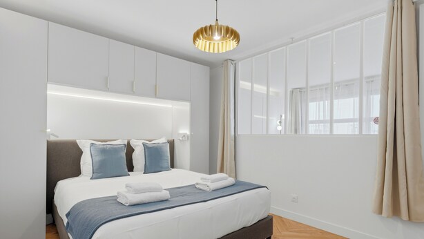 Une chambre d’hôtel moderne avec un grand lit, une armoire intégrée, une grande fenêtre avec des rideaux et un luminaire suspendu.