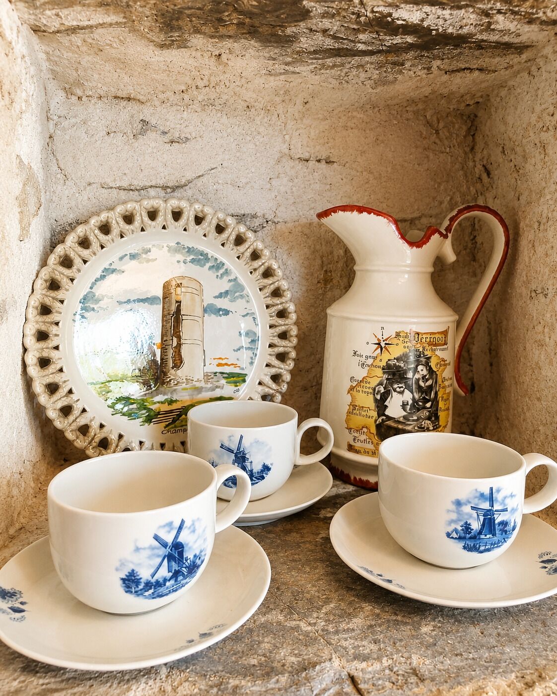 A set of white ceramic cups and saucers with blue windmill designs, a decorative plate with a similar motif, and a white pitcher with red trim and a windmill illustration.