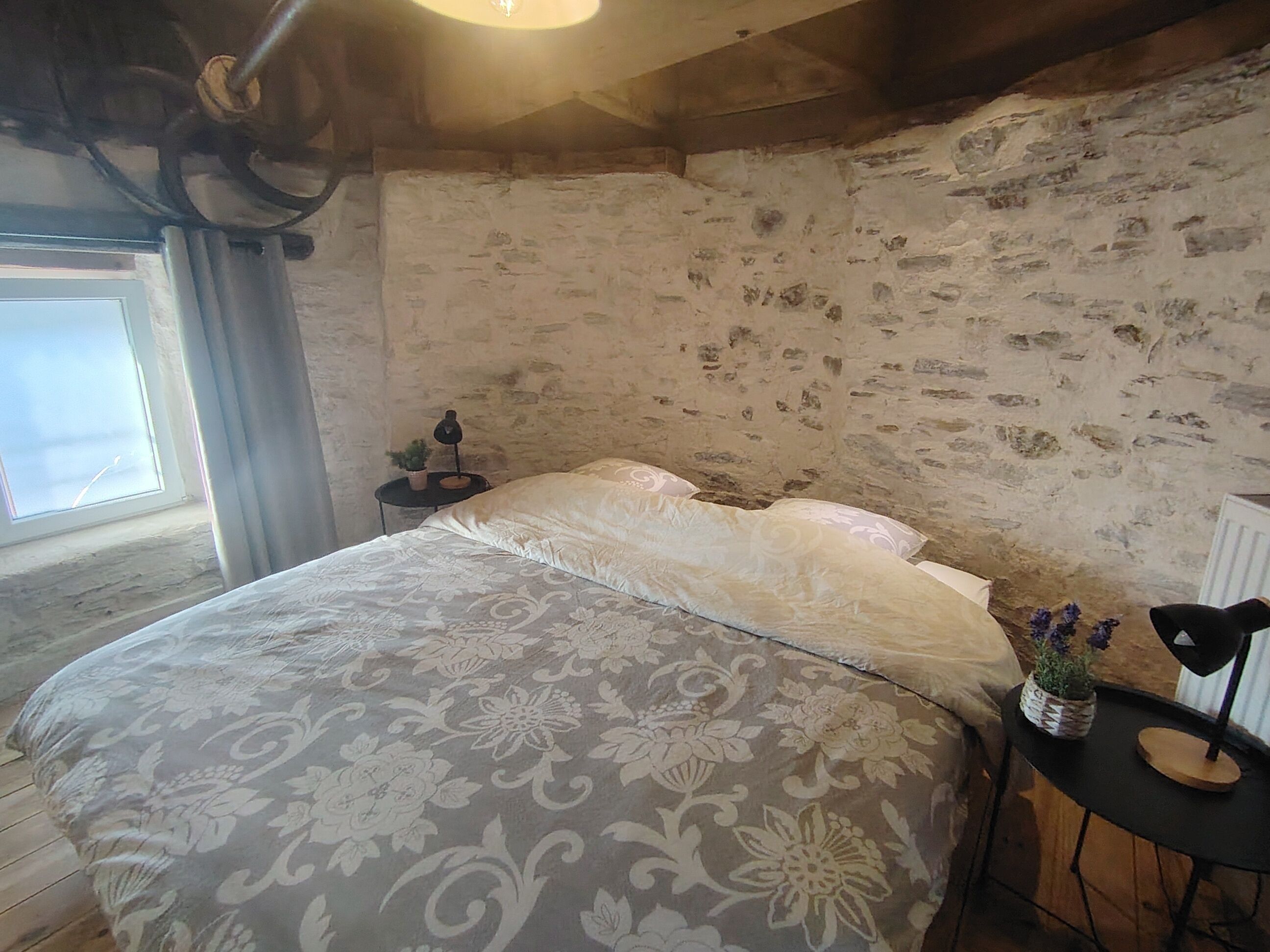 A bedroom with a bed, a small bedside table with a lamp, a potted plant, and a window with curtains.