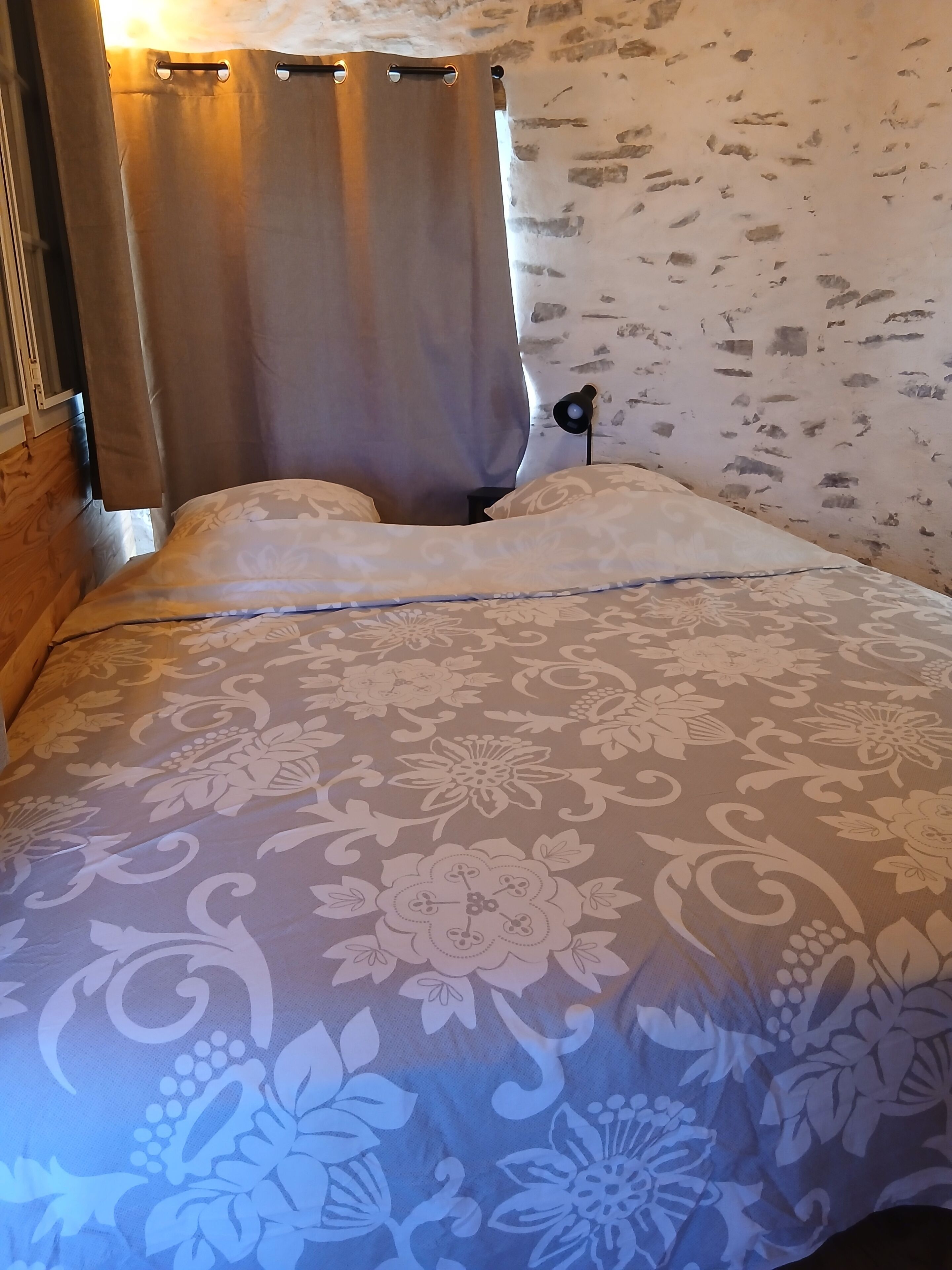 Ein Bett mit floralem Überwurf, ein Kopfteil mit Lichtschalter und ein teilweise gezogener Vorhang.