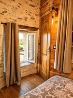 Um quarto com porta de madeira, janela com cortinas, cama com colcha estampada e luminária sobre a porta.