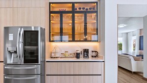 Uma cozinha moderna com geladeira de aço inoxidável, um armário com porta de vidro e utensílios de cozinha, e bancada com cafeteira.