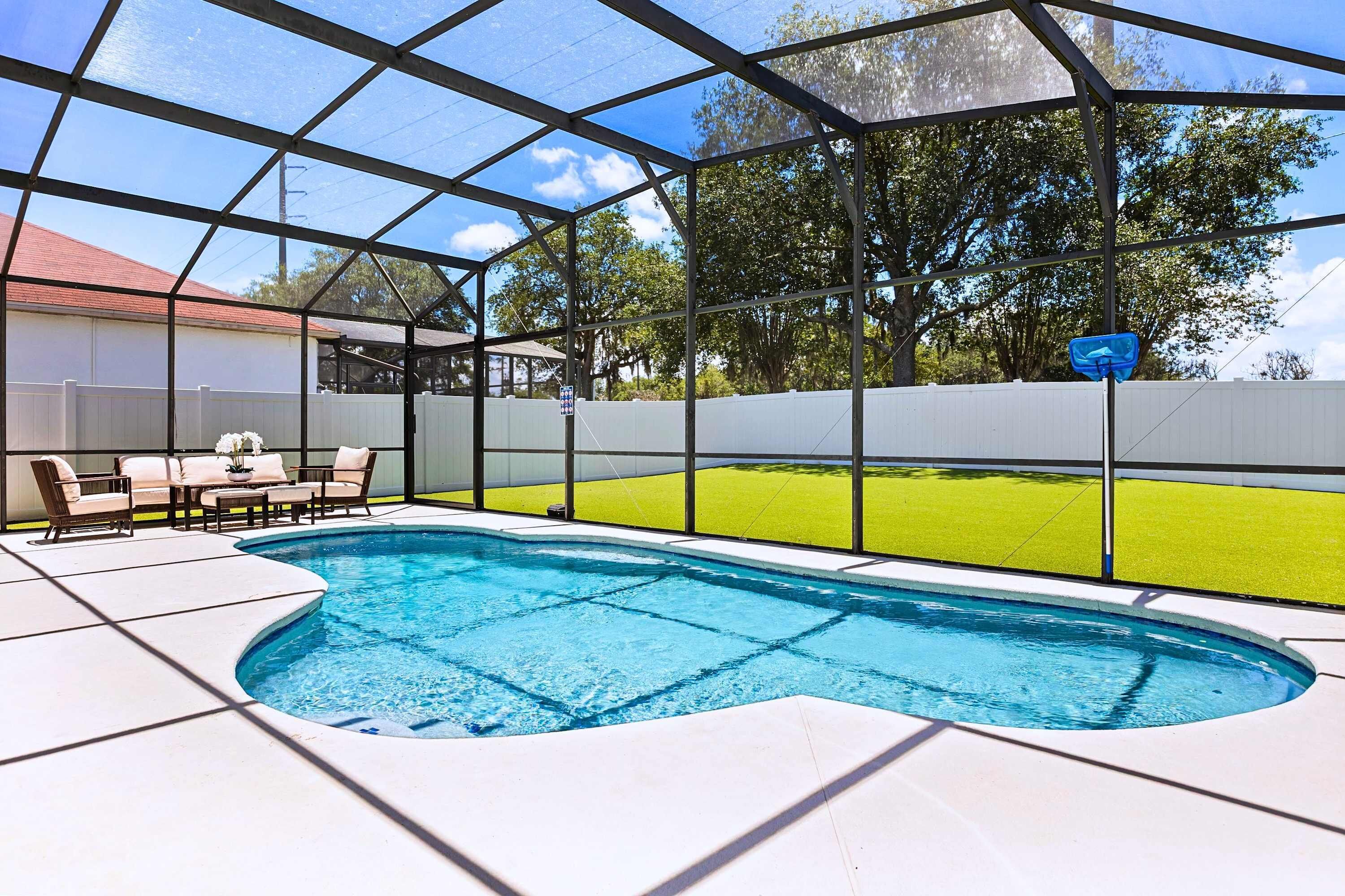 Una piscina con zona cubierta, un patio con sillas y vistas a un césped verde.