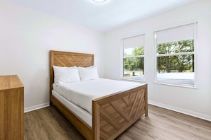 A bedroom with a wooden bed, white bedding, and two windows with a view of trees. - Upstay Contemporary Retreat w Pool, BBQ & Backyard (Davenport)