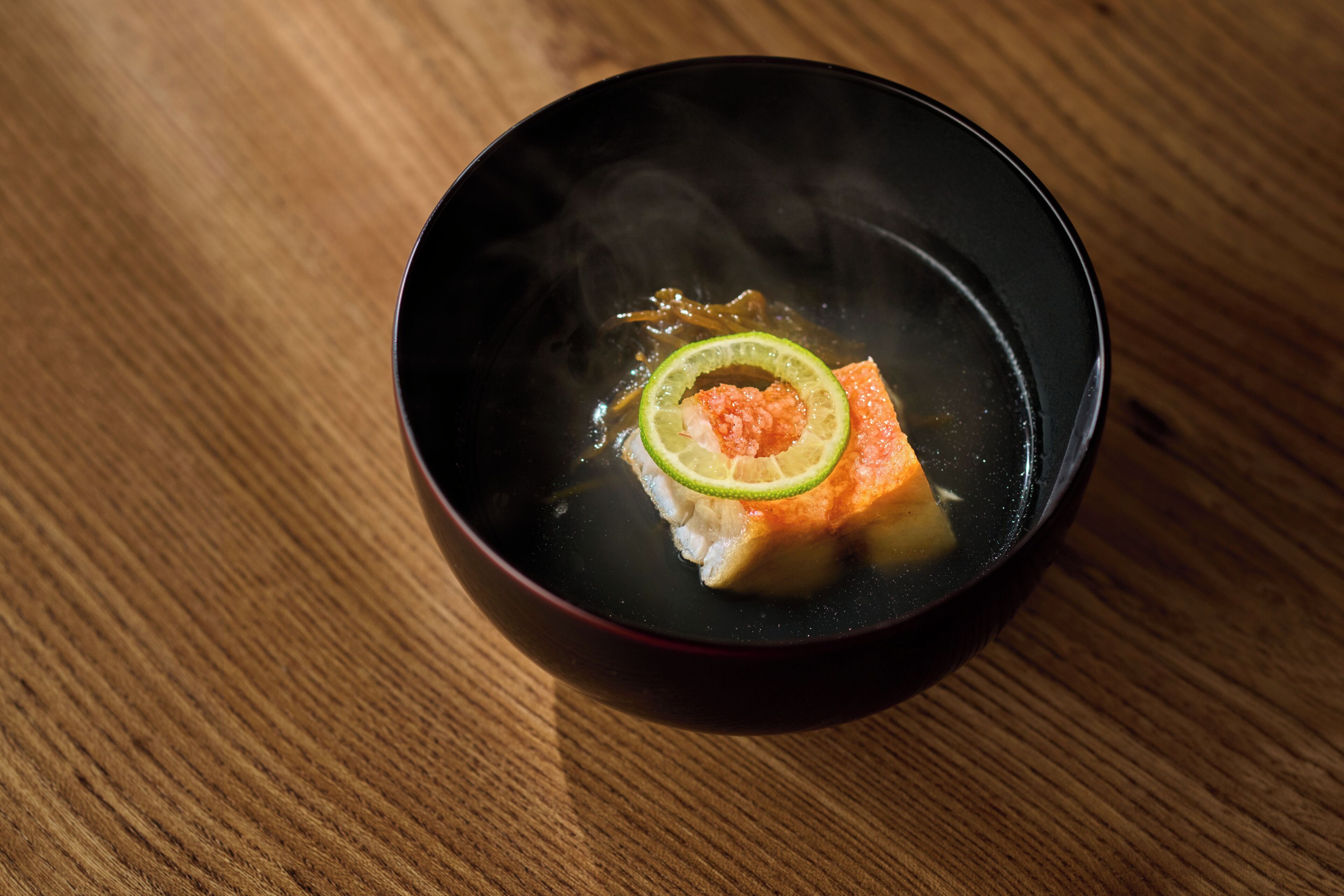 Un bol noir contenant un morceau de saumon et une tranche de citron vert, posé sur une table en bois.
