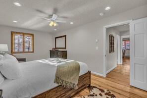 Quarto com cama, ventilador de teto, cômoda, espelho e porta que dá para outro ambiente.