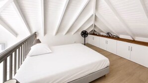 Een slaapkamer met een schuine plafond, een bed, een ventilator en een houten plank met boeken.