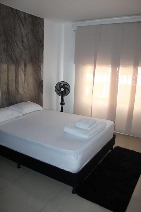 Un dormitorio con cama, ventilador de pie, ventana con cortinas y alfombra oscura.