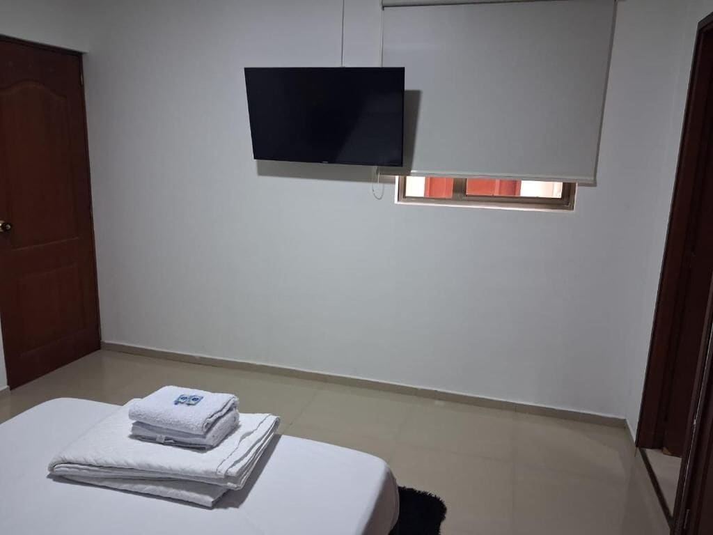 Una habitación con una cama, un televisor de pantalla plana montado en la pared y una ventana con persianas.