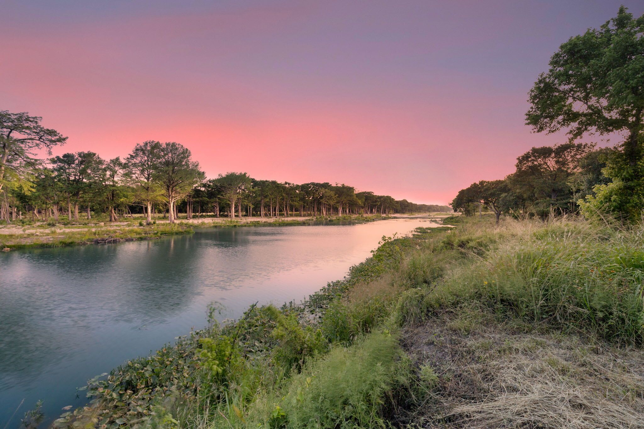 Une rivière bordée d’arbres des deux côtés, un ciel dégagé et une surface d’eau calme.