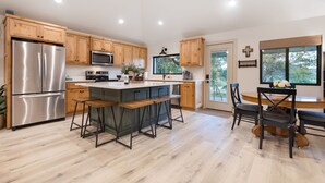 A modern kitchen with wooden cabinets, stainless steel appliances, a central island with bar stools, and a dining area with a wooden table and chairs. - Twisted Cedar Retreat - River views w/Firepit! (Ingram)