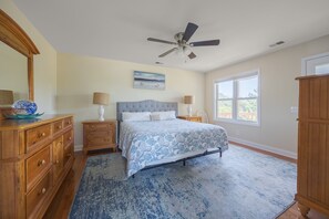 Ein Schlafzimmer mit einem Bett, Nachttischen, einer Kommode, einem Deckenventilator, einem Fenster und einem Bild an der Wand.