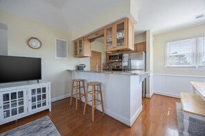 A compact kitchen with wooden cabinets, a built-in microwave, and a refrigerator. There are two stools and a television in the adjacent living area. - 5600 - On the Sunny Side Soundside 3BR retreat with deck, Wi-Fi, and golf views. (Nags Head)