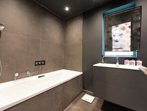 A modern bathroom with a large white bathtub, a mirror with a blue frame, and a small shelf with toiletries. - Architect-Designed Luxury Retreat, 3BR Chessington (Chessington)