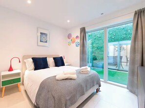 A modern bedroom with a large bed, a bedside table, a green drawer, and a view of a garden through a sliding glass door. - Architect-Designed Luxury Retreat, 3BR Chessington (Chessington)