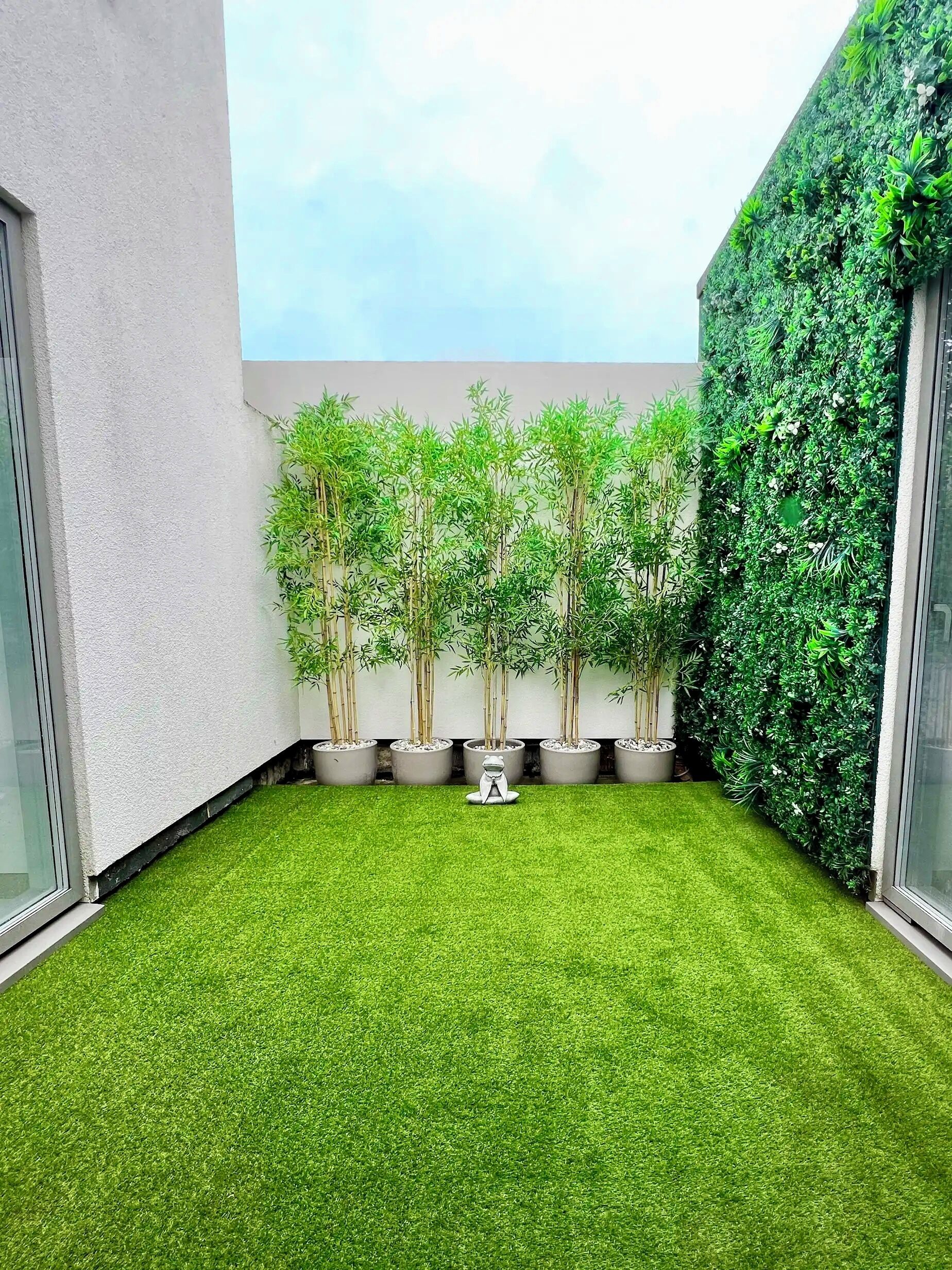 Un patio con césped artificial, plantas de bambú en macetas y un jardín vertical verde.