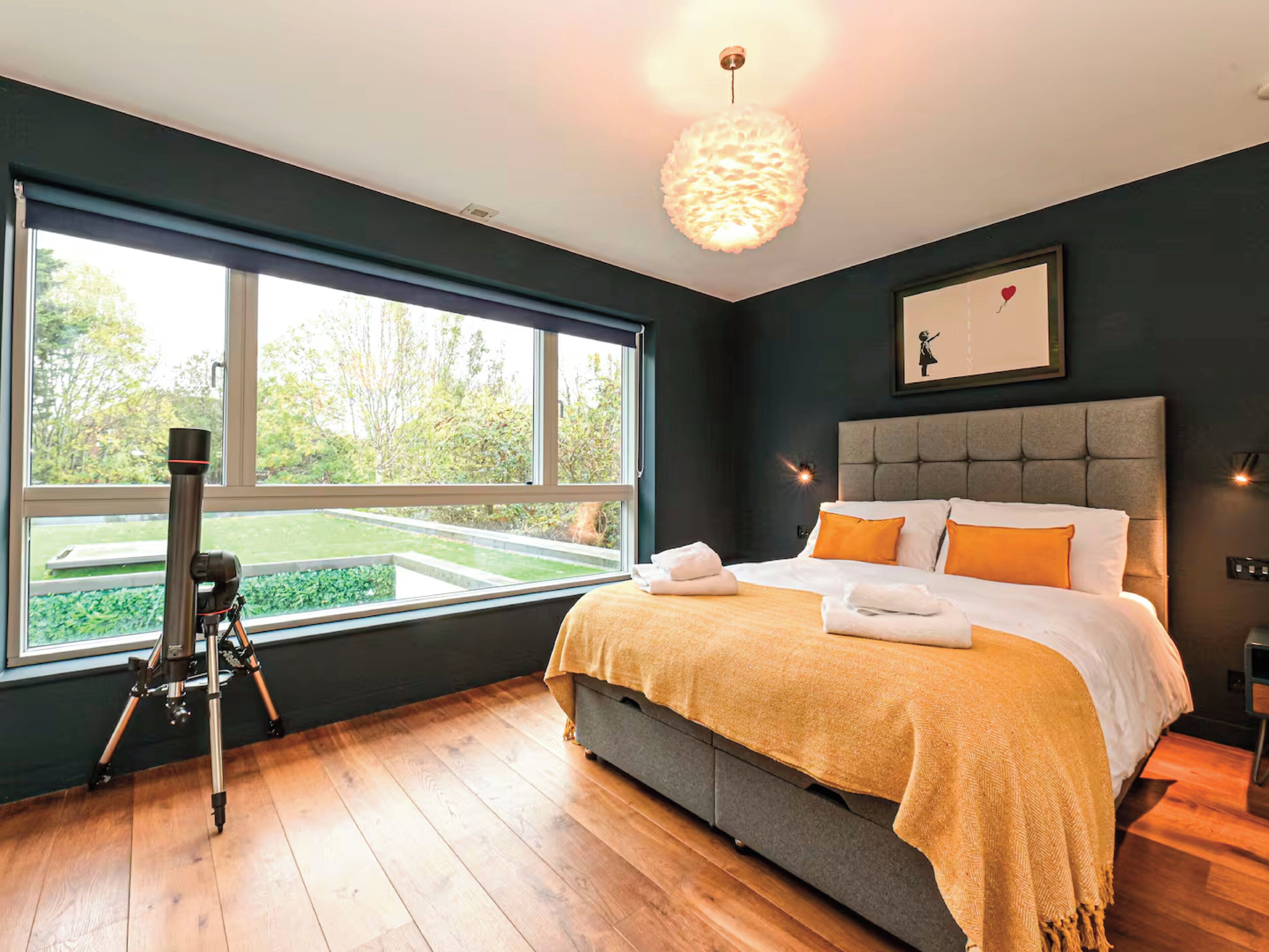 A bedroom with a large window, a telescope, a bed with orange pillows, and a framed picture on the wall.
