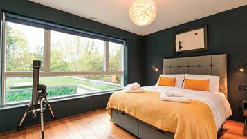 A bedroom with a large window, a telescope, a bed with orange pillows, and a framed picture on the wall.