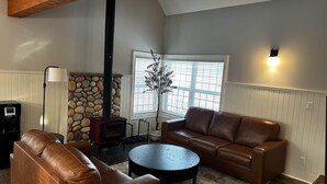 A living room with a brown leather sofa, a round coffee table, a stone fireplace, and a large window with blinds. - Charming  Rustic cabin w/Jacuzzi at Fowler Lake, SK (Loon Lake No. 561)