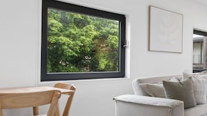 A room with a large window, a dining table with chairs, and a sofa with cushions. - Sandy Calm, Manly Walk to Beach, Corso & Ferry (Manly)